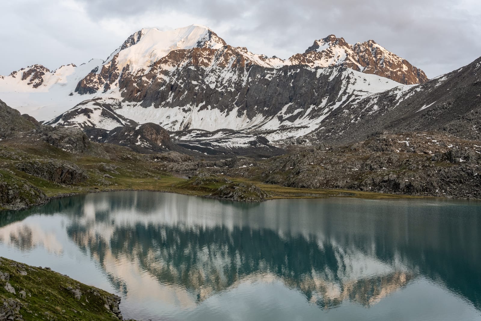 Snowy peaks in warm light mirrored in a still alpine lake under soft cloud