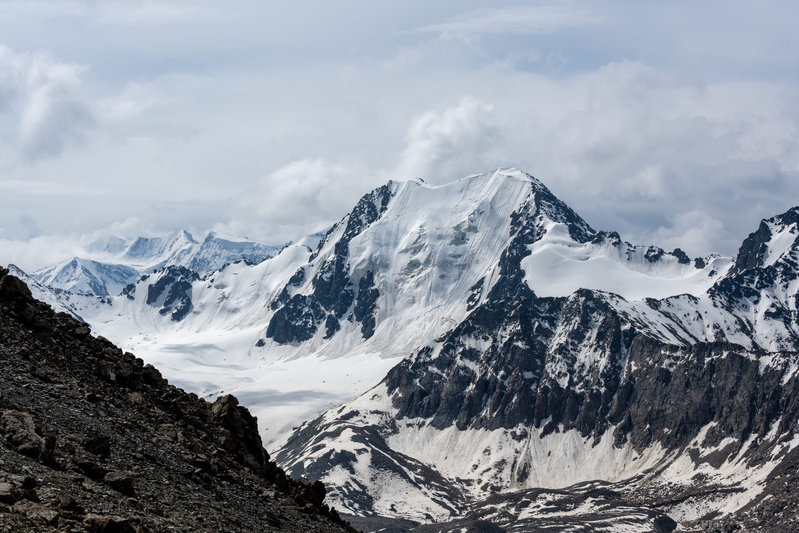 Glaciated white summit and icefield seen from a high pass with a dark rock foreground