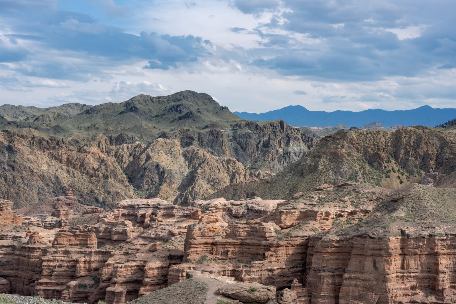 Red rock canyon towers below folded grey-green mountains under broken cloud