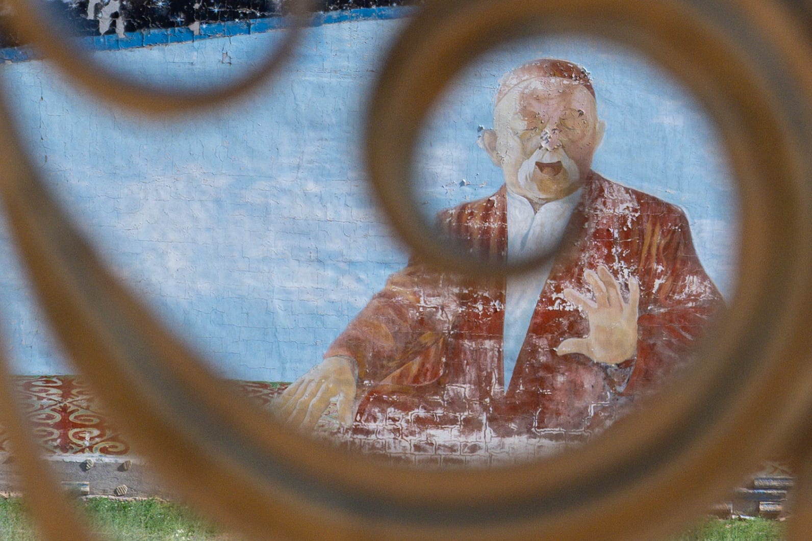 Weathered mural of a bearded man seen through a rusted iron scroll