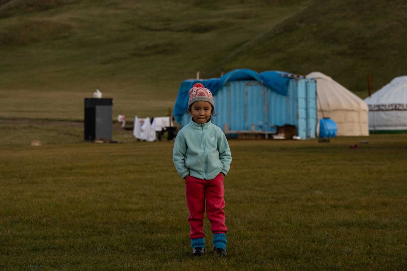 Small child in a pink striped hat, pale green fleece and red trousers standing on a meadow, yurts and a blue shipping container behind