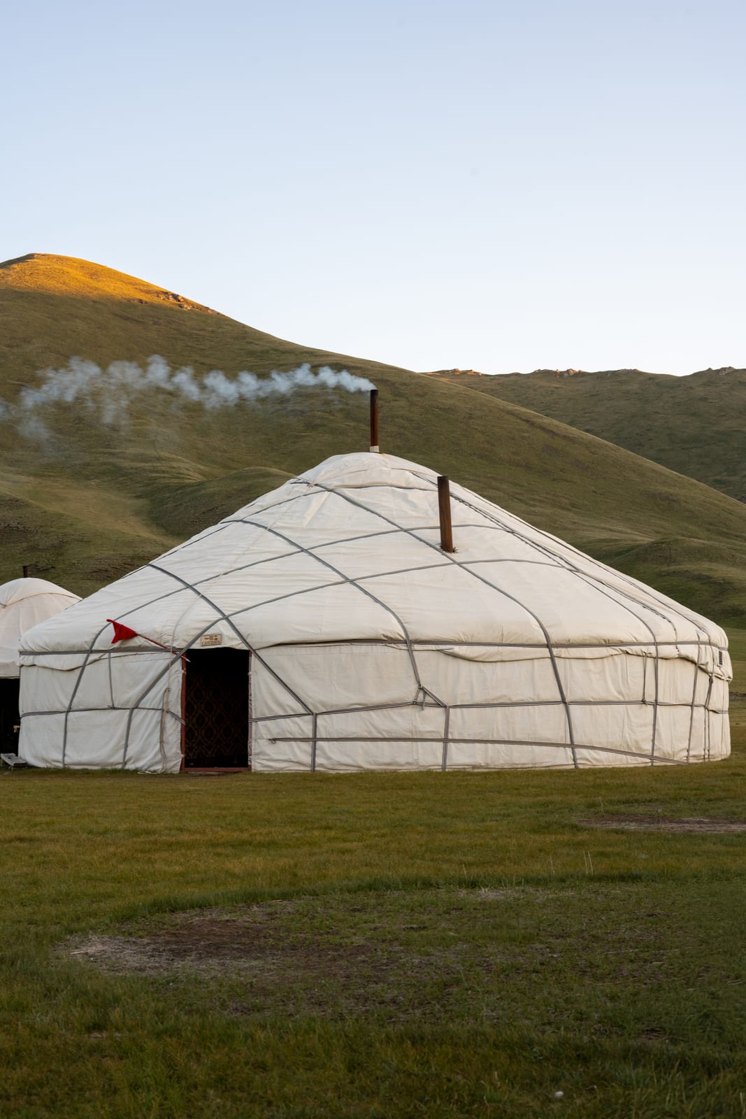 White yurt with chimney smoke drifting across a green hillside at evening