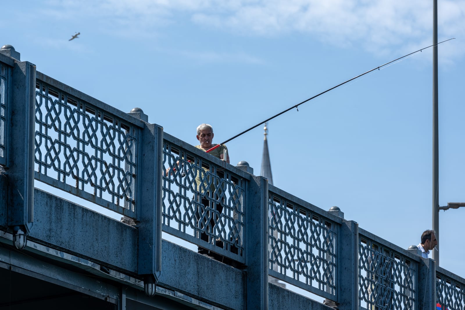 Fisherman leaning over the blue lattice railing of Galata Bridge, rod cutting diagonally across the sky