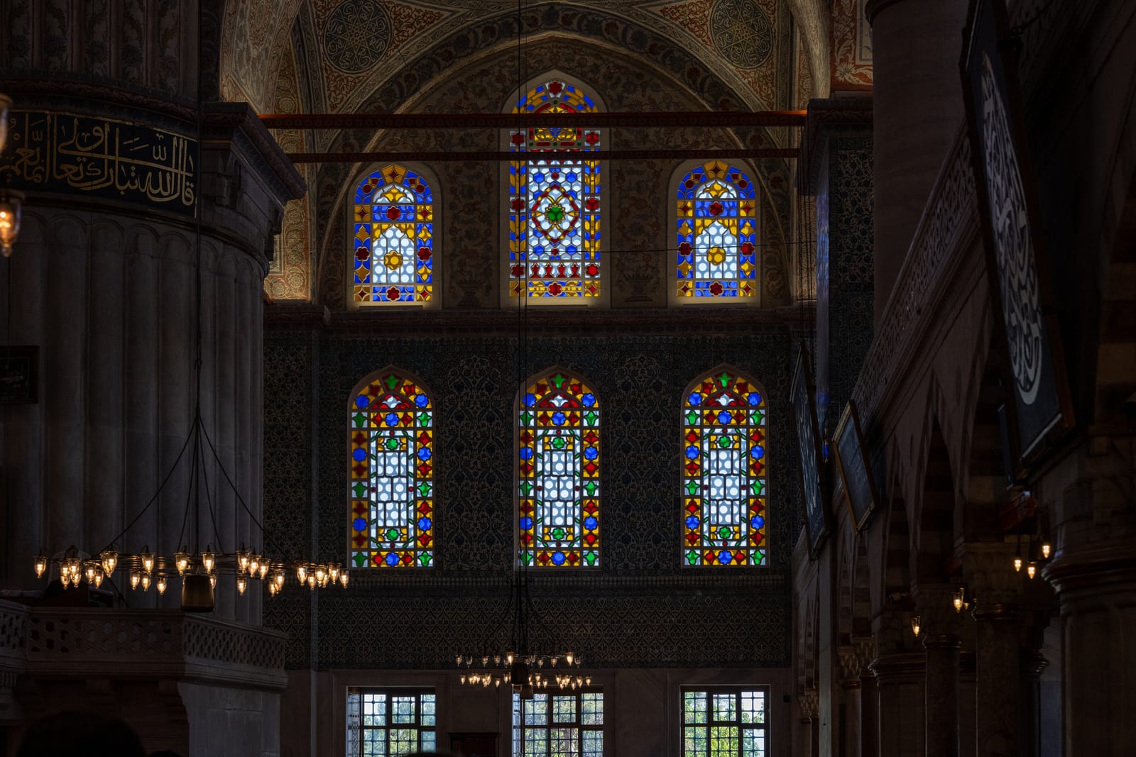 Dark mosque interior with two tiers of glowing stained-glass windows and lamp rings