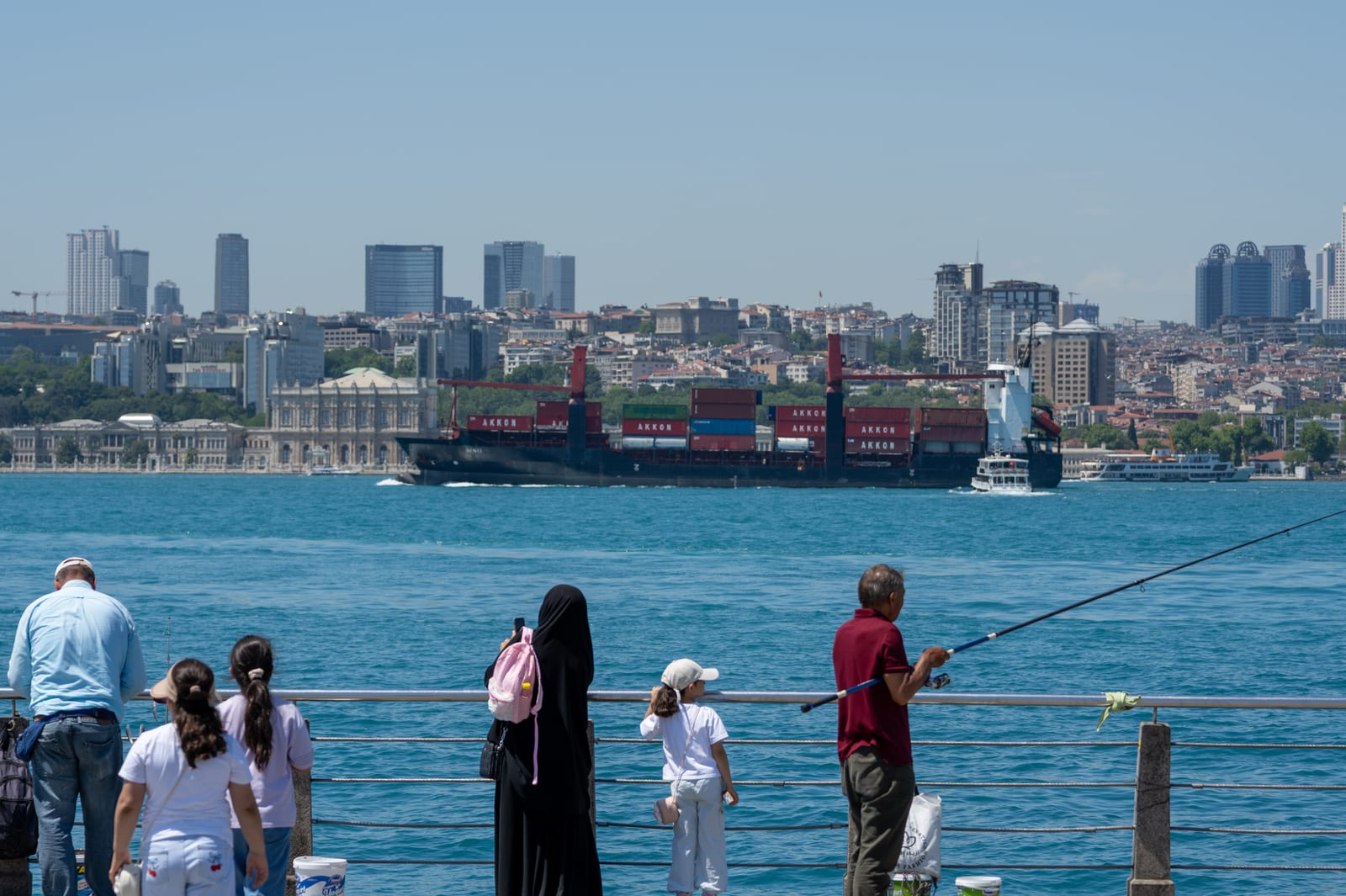 Row of anglers and passers-by on the Bosphorus waterfront, container ship and skyline behind