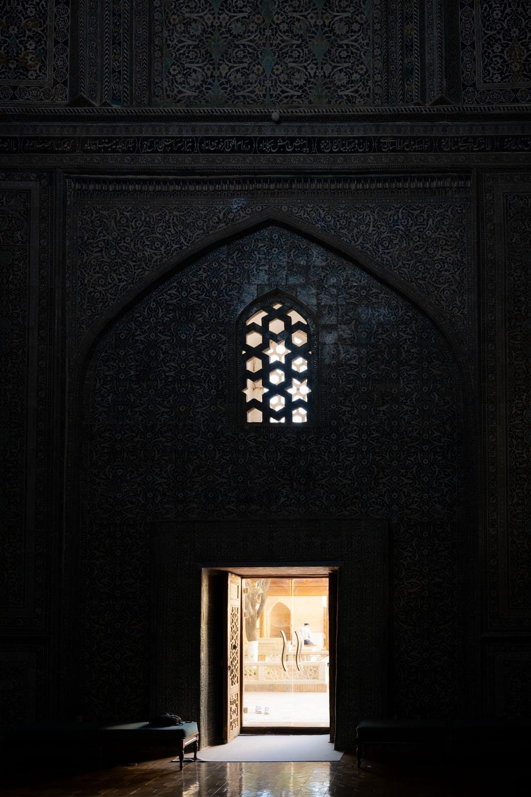 Near-black interior with a latticed window and a small bright doorway opening to daylight