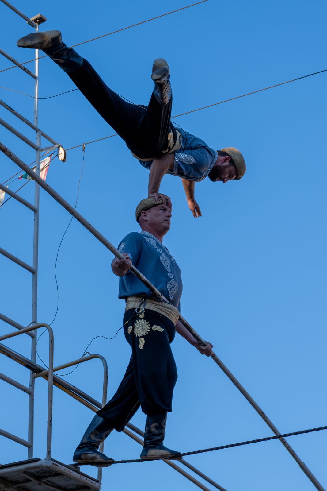 Tightrope walker with balance pole carrying a second performer doing a handstand on his shoulders, blue sky