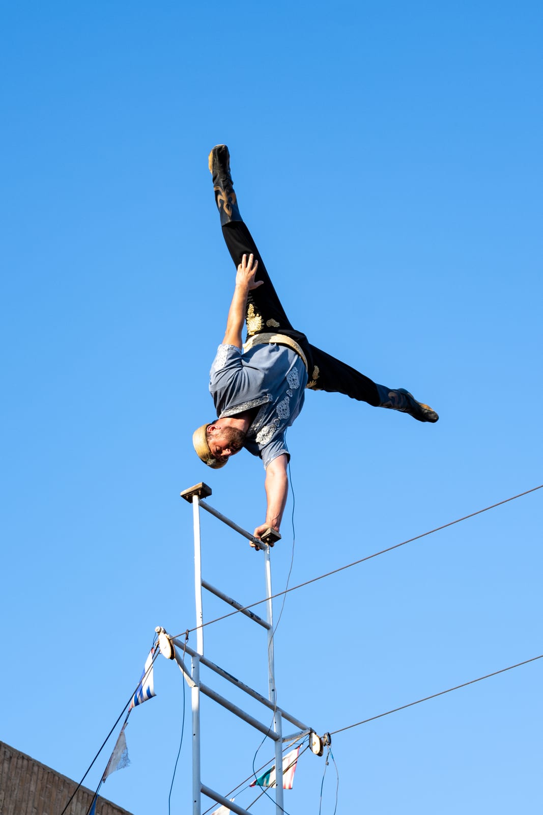 Acrobat in a one-arm handstand split on top of a mast against a clean blue sky