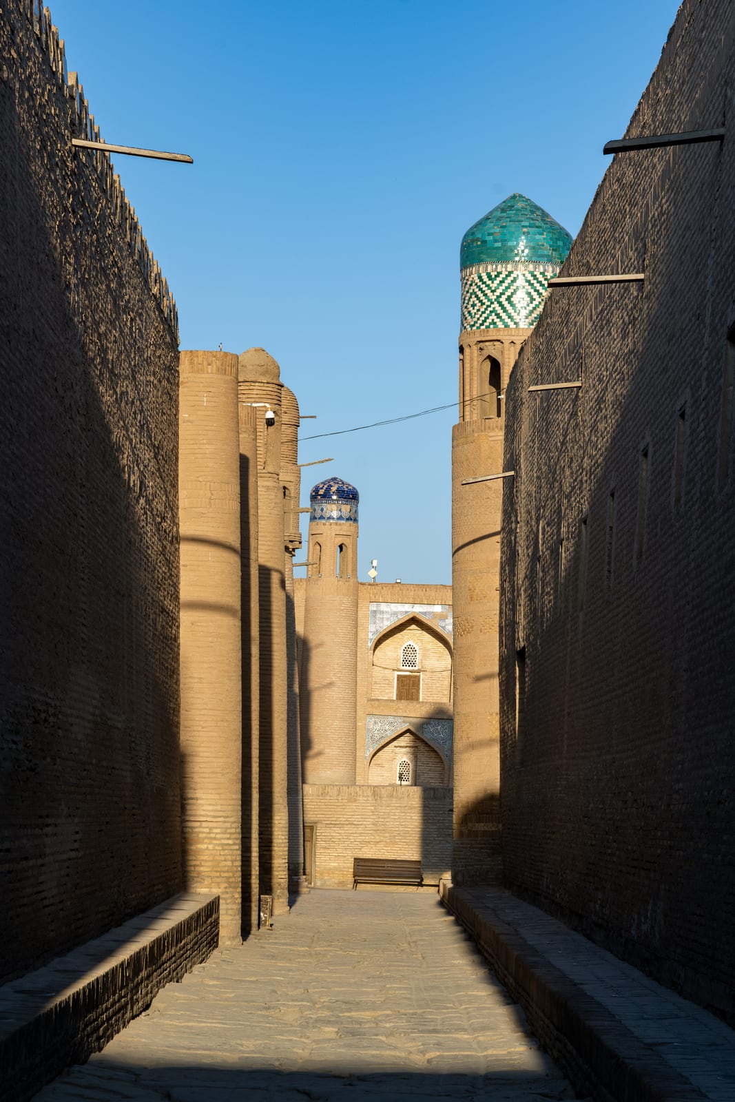Narrow shadowed alley between high mud-brick walls opening onto a tiled minaret