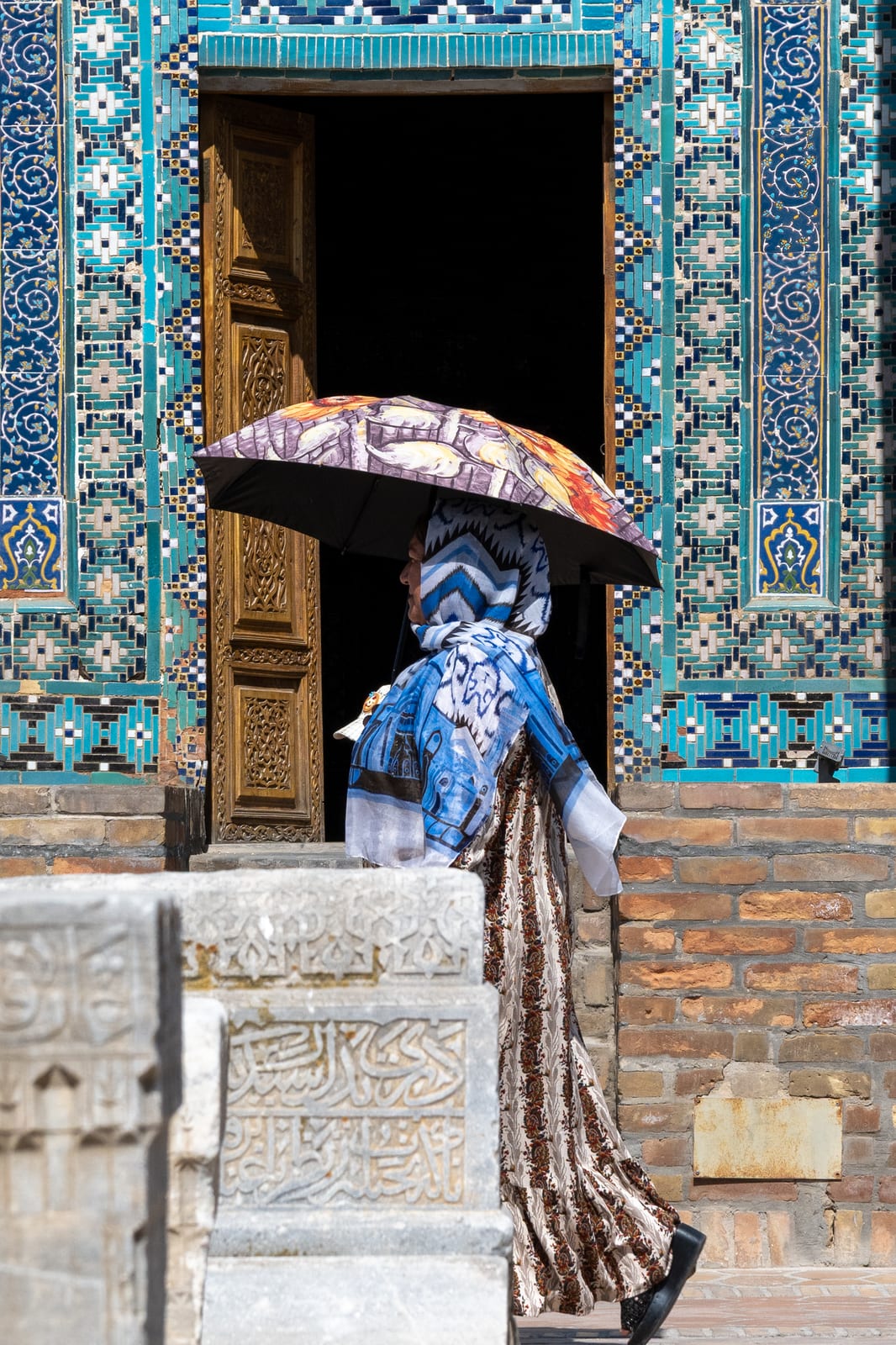 Woman in a blue headscarf with a patterned parasol walking past a turquoise-tiled mausoleum doorway