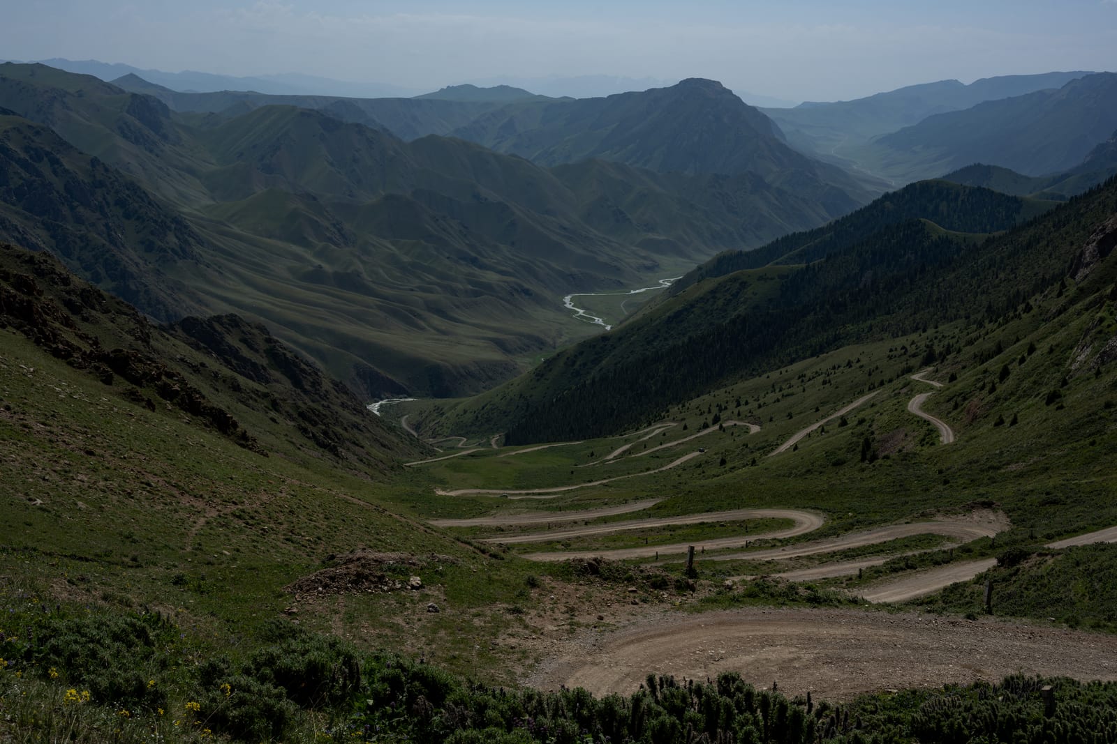 Dirt road switchbacking down a green mountain pass into layered hazy valleys
