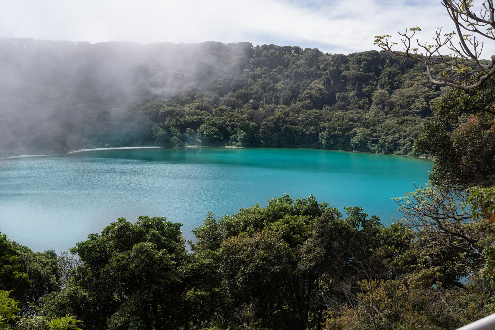 Turquoise crater lagoon ringed by forest with mist drifting across the water
