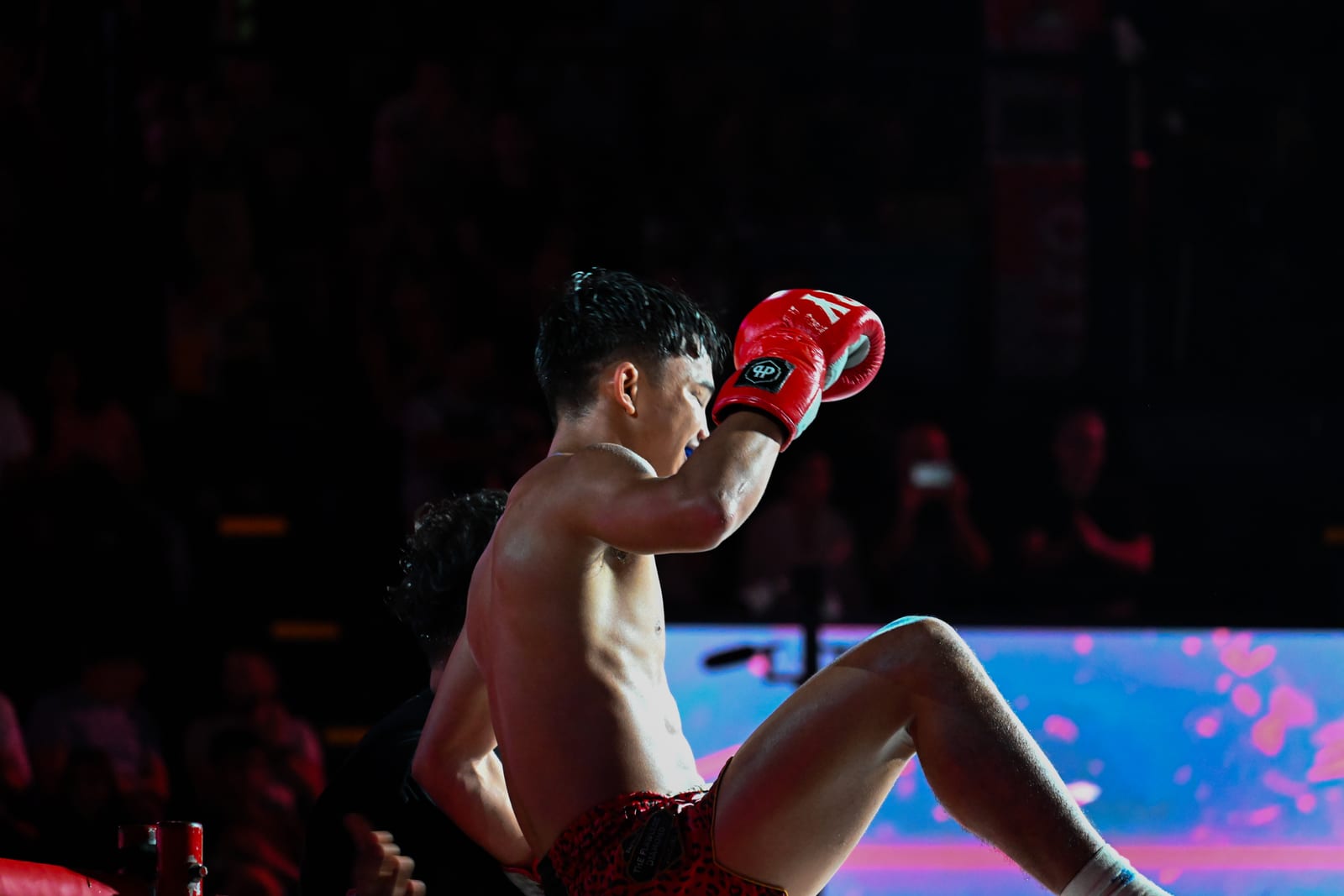 Muay Thai fighter seated on the canvas with glove raised, dark arena