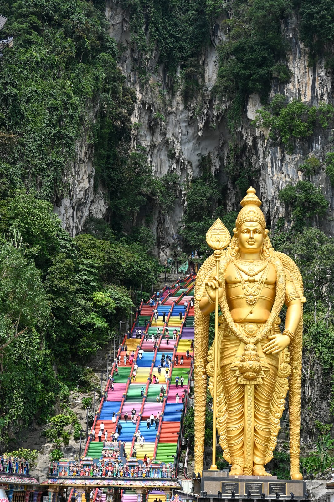 Full Murugan statue beside the rainbow staircase beneath jungle cliffs