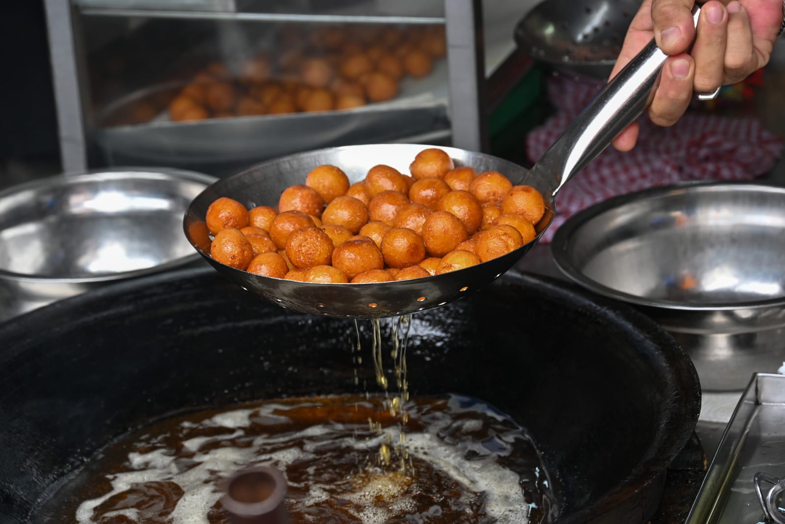 Hand lifting a strainer of fried dough balls over a wok of hot oil