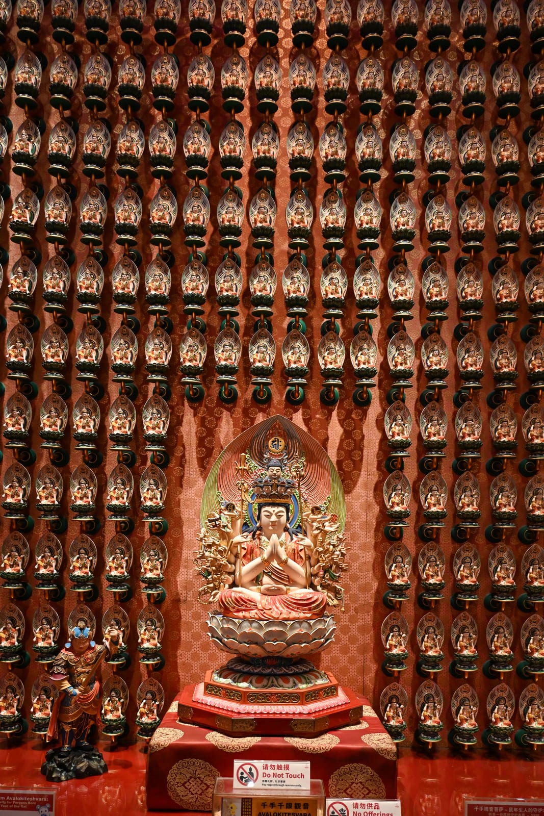 Wall of hundreds of small Buddha figures behind a seated deity, red and gold