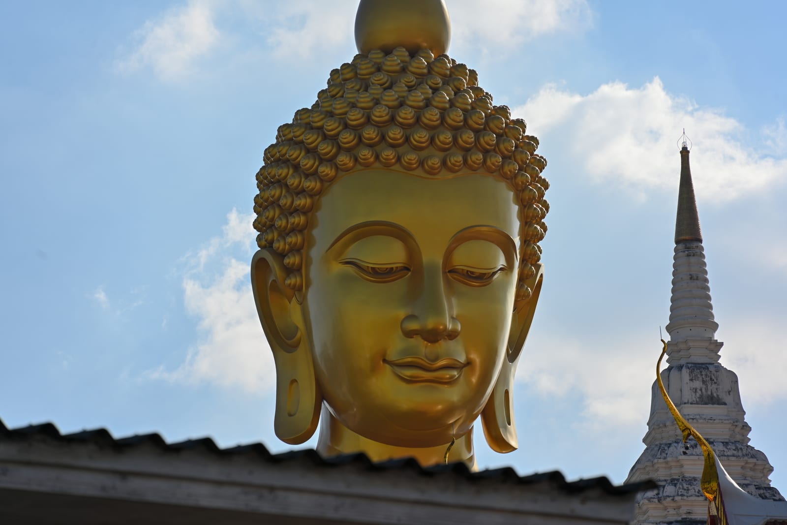Giant golden Buddha head rising above a rooftop against blue sky and clouds, white spire at right