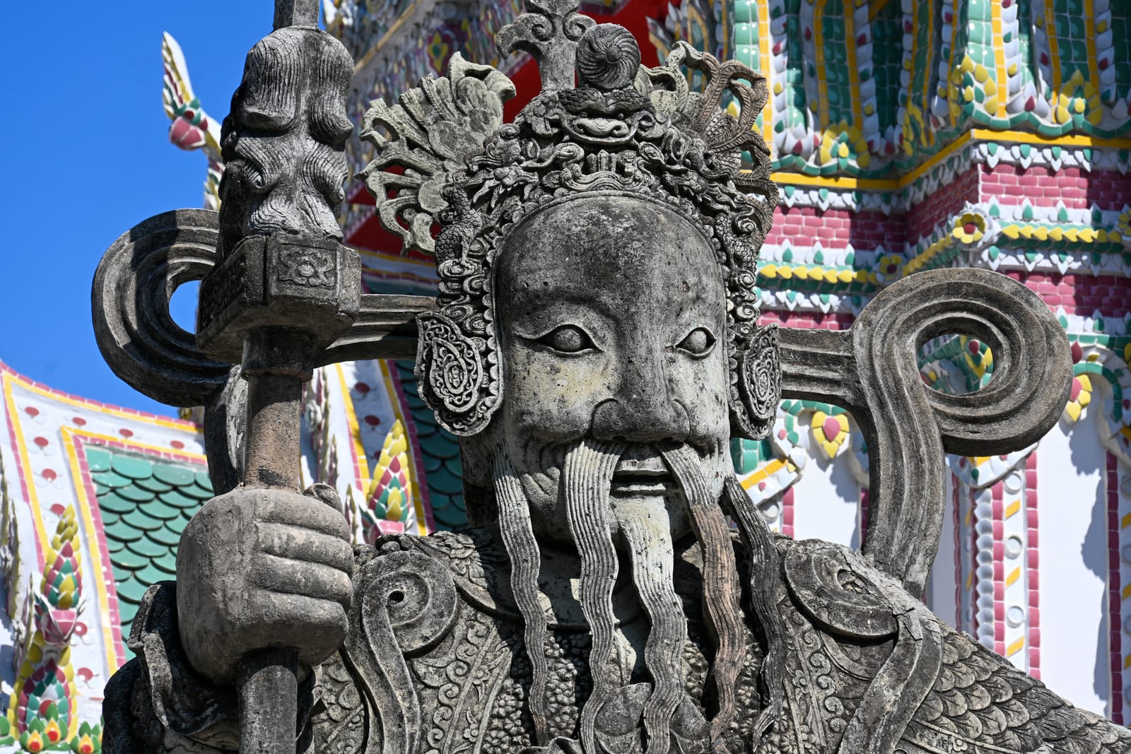 Stone Chinese guardian statue face with long beard, colourful tiled chedi behind