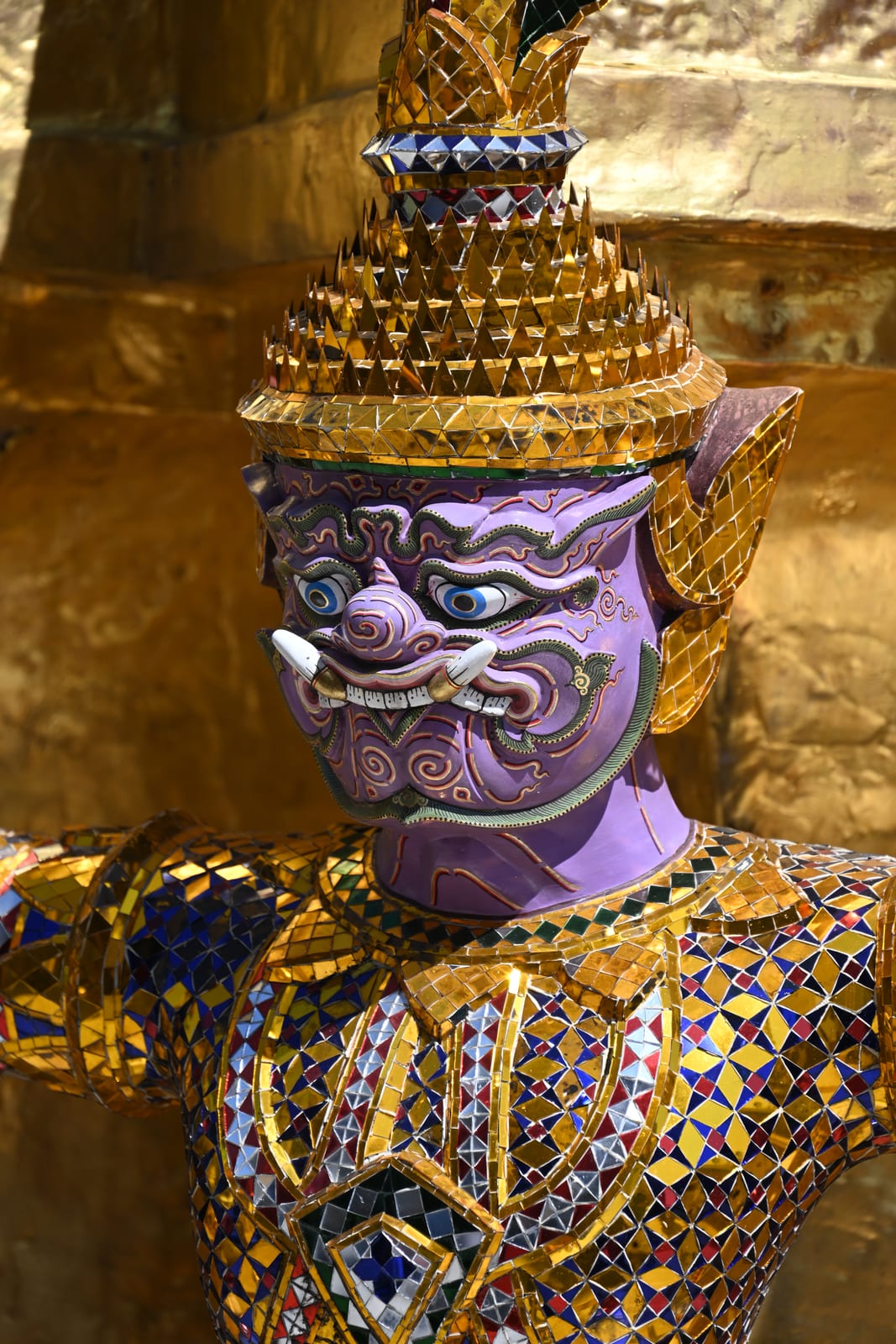 Purple-faced demon guardian figure in jewelled gold armour against a gold wall at the Grand Palace