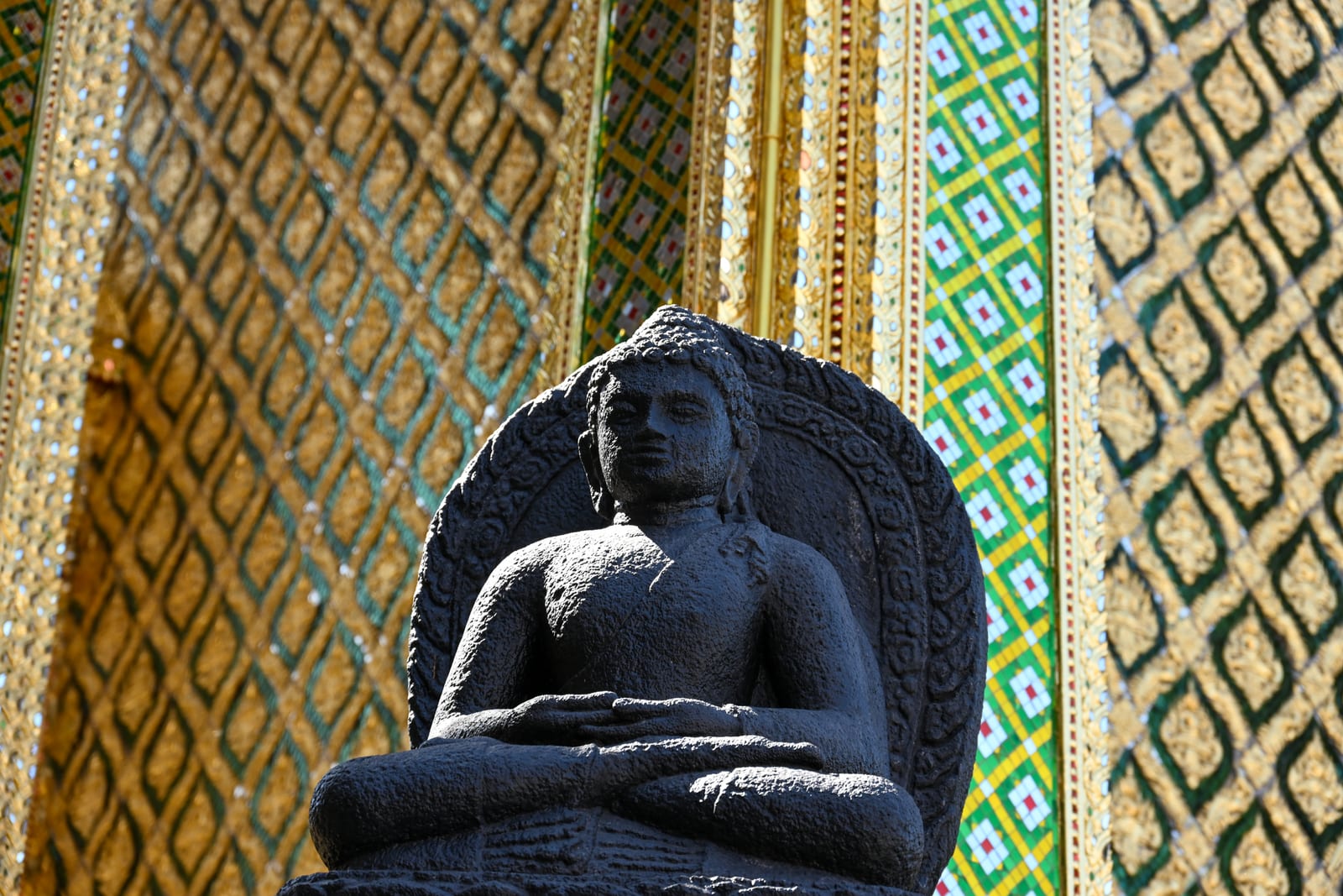 Dark stone seated Buddha against patterned gold and green mosaic columns