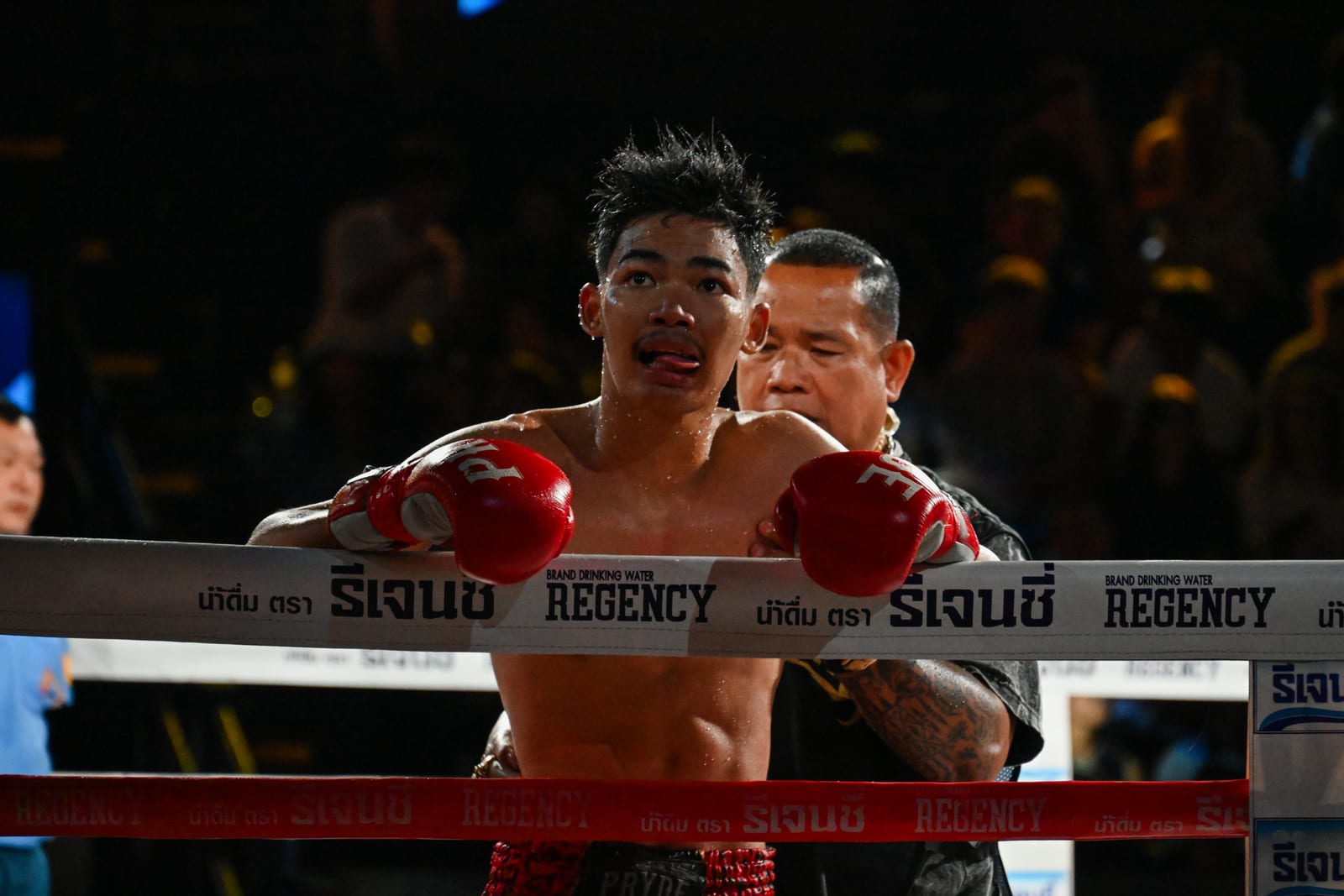 Muay Thai fighter leaning on the ropes with red gloves, cornerman behind