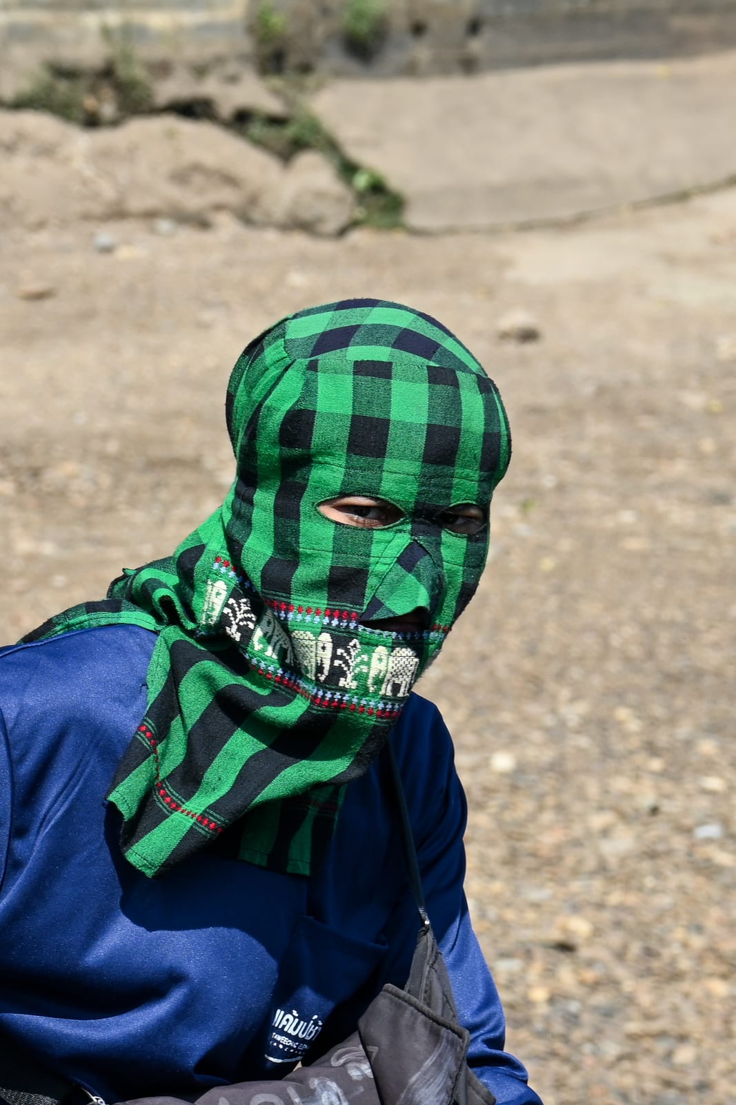 Mahout in a green checked face covering and blue jacket, sandy background