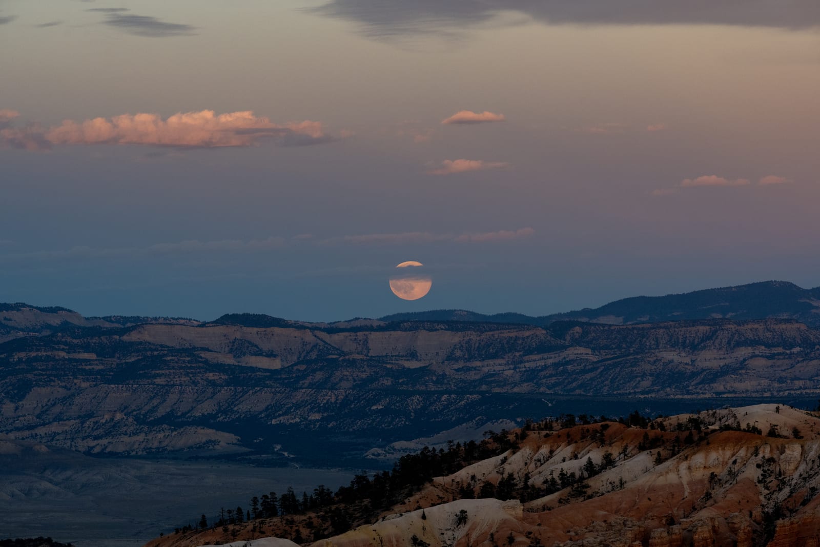 Full moon rising through dusk haze over layered blue ridges, pink clouds, hoodoo slopes in foreground
